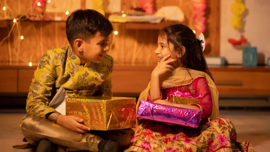 Two children in traditional Indian outfits smiling and exchanging wrapped gifts at Diwali, with fairy lights in the background.